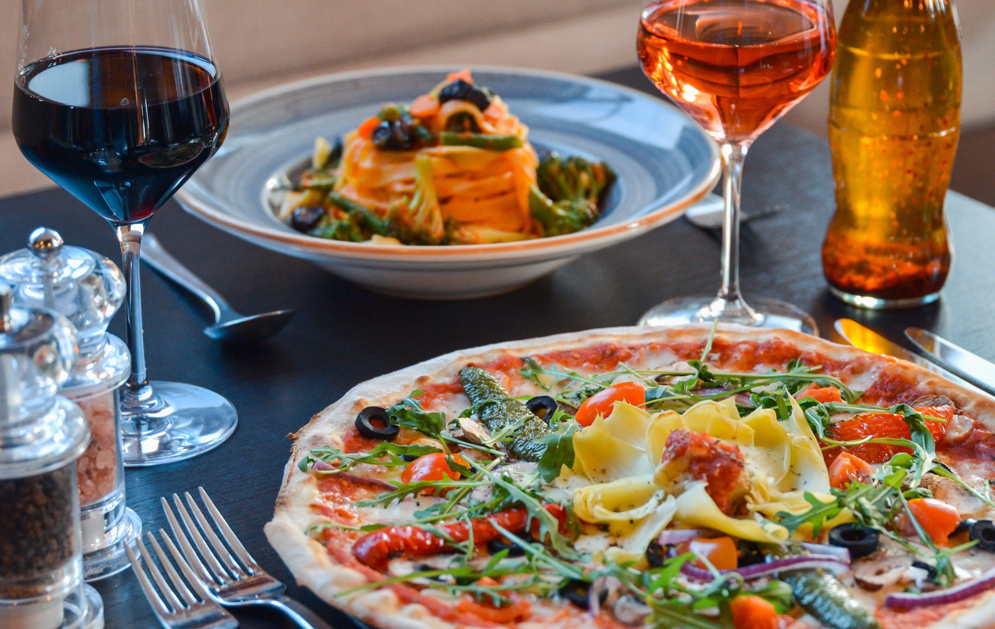 A pizza, a pasta dish and two glasses of wine on a restaurant table