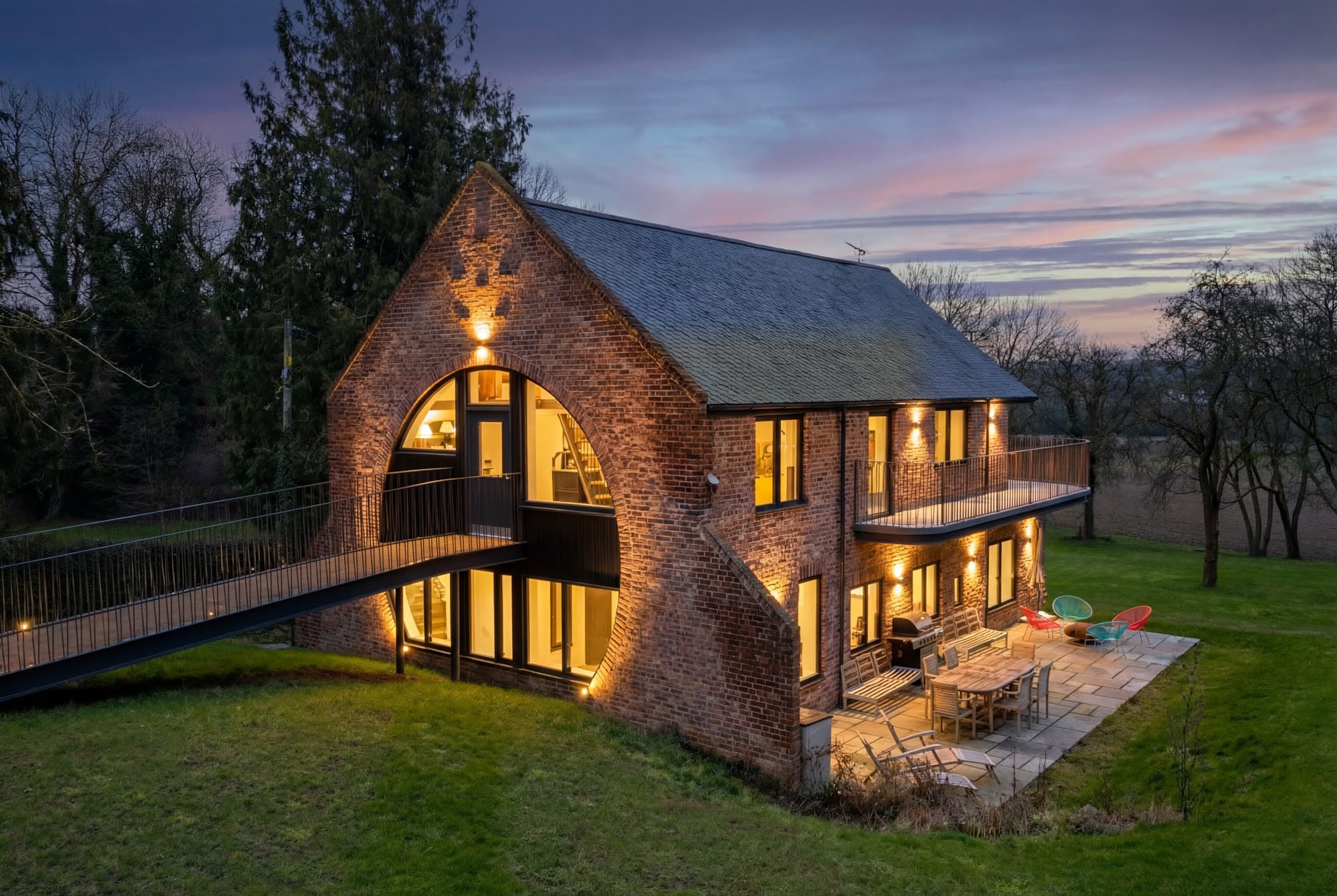 A brick barn house at twilight with an arched glass gable, a footbridge and a lit terrace